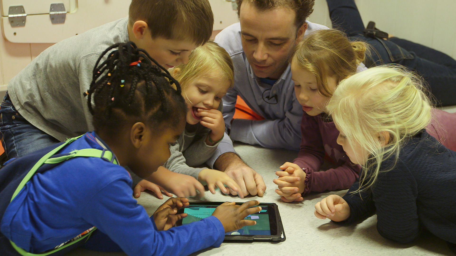 Barnehagelærer og fem barn ligger rundt et nettbrett på gulvet