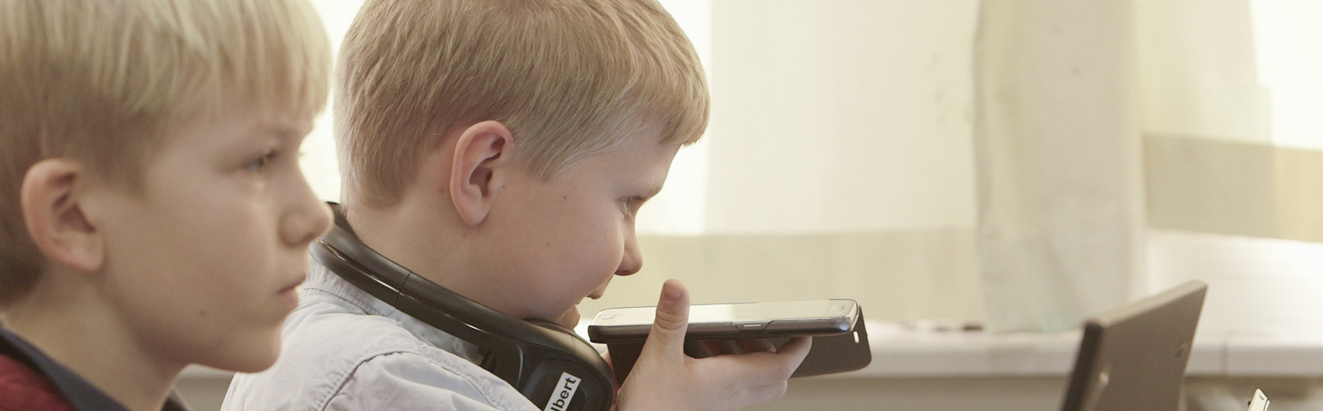En gutt snakker inn i en telefon han holder flatt foran seg. En annen gutt ser på en skjerm.